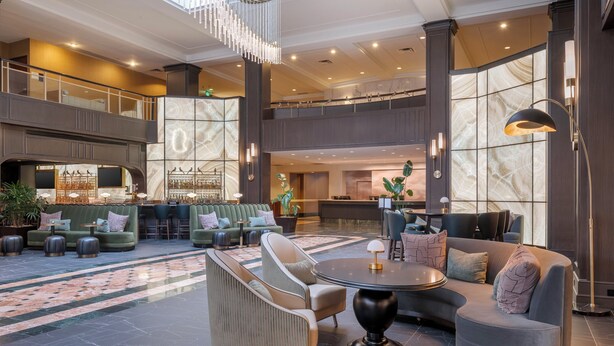 Un lobby amplio con techo alto, un área de estar central con sofás y sillones, una zona de bar con mostrador y taburetes, y una gran pared decorativa con un diseño geométrico.