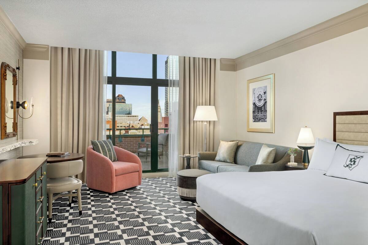 A hotel room with a large bed, a desk, a chair, a sofa, and a view of the cityscape through the window.