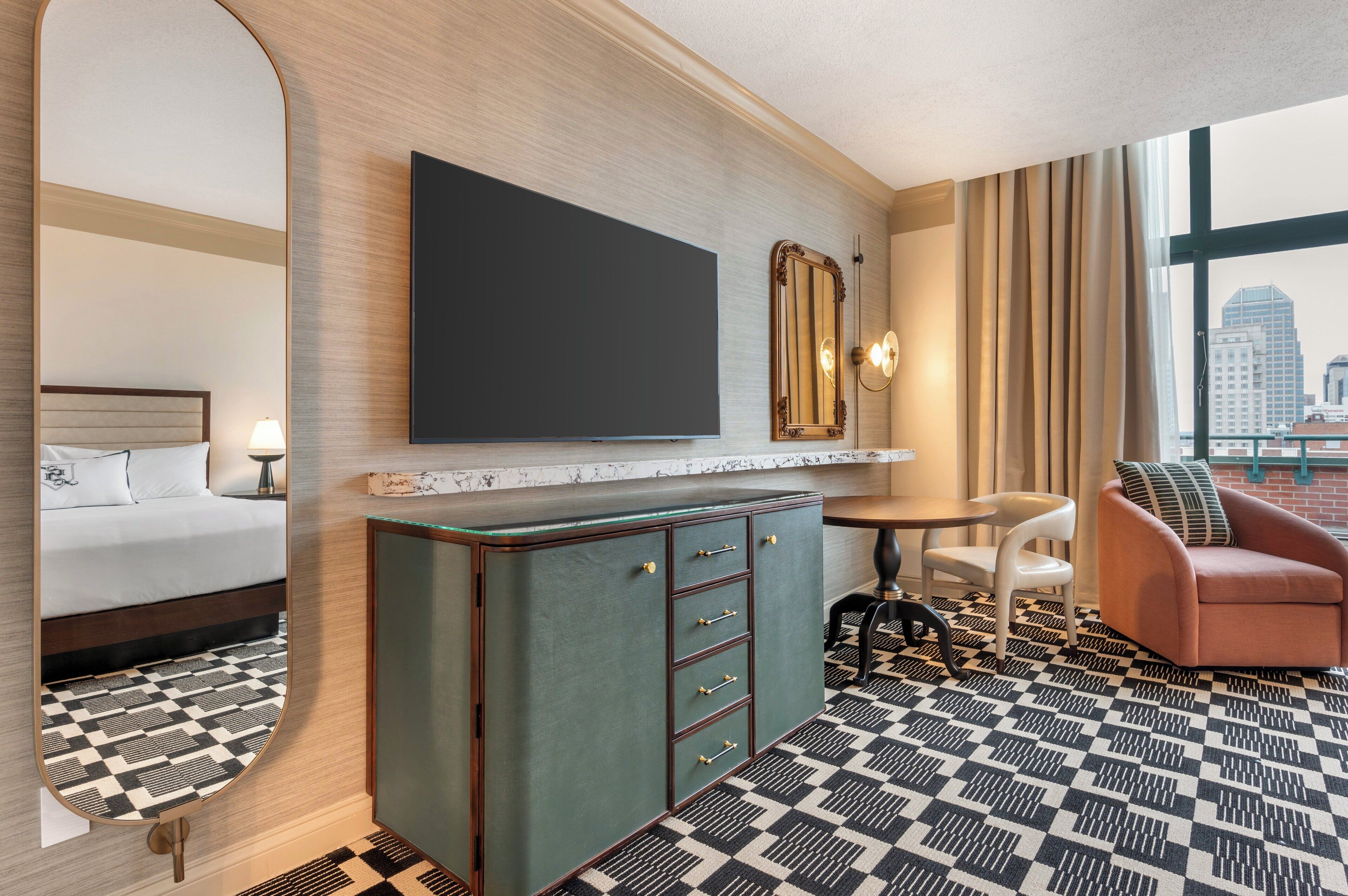 Habitación de hotel con cama, minibar, televisión, espejo, zona de estar con una silla y un sofá, y piso estampado.