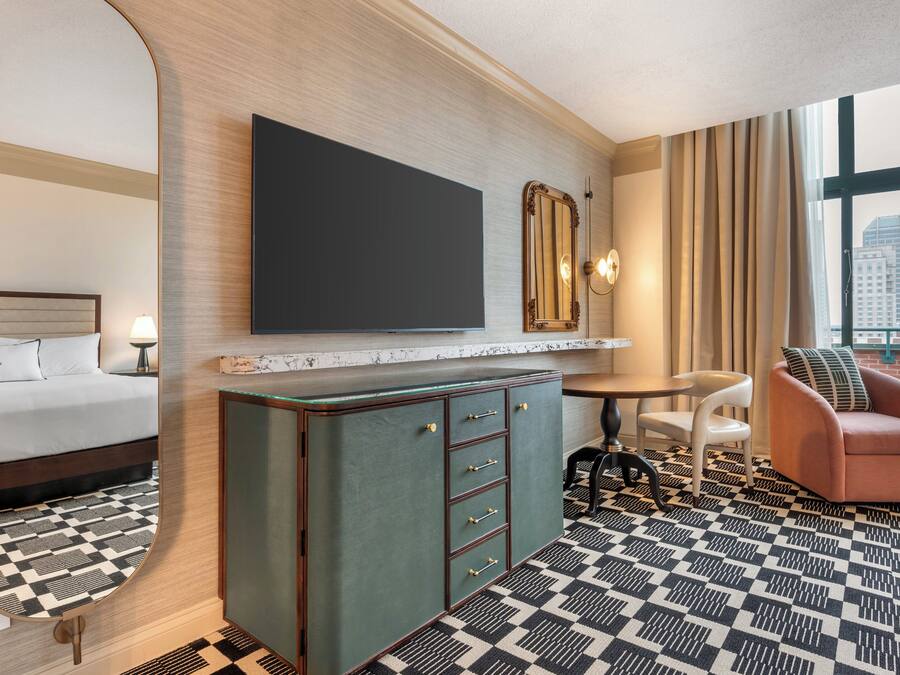 Habitación de hotel con cama, minibar, televisión, espejo, zona de estar con una silla y un sofá, y piso estampado.