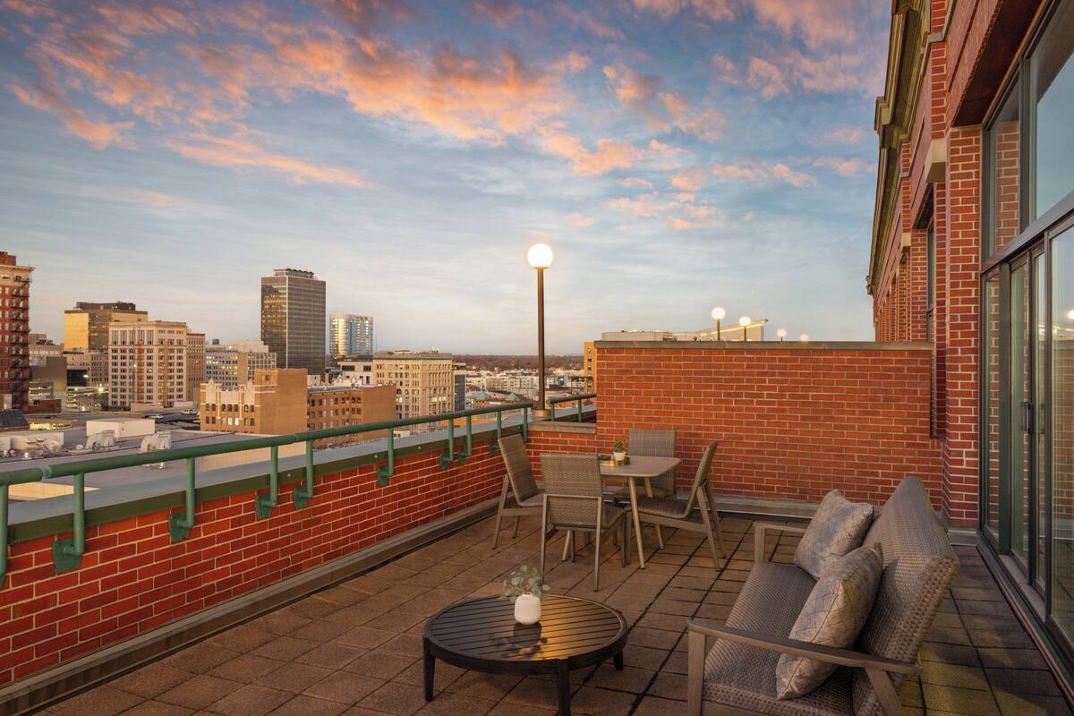 A rooftop terrace with a table, chairs, and a sofa, overlooking a cityscape with buildings and a sunset sky.
