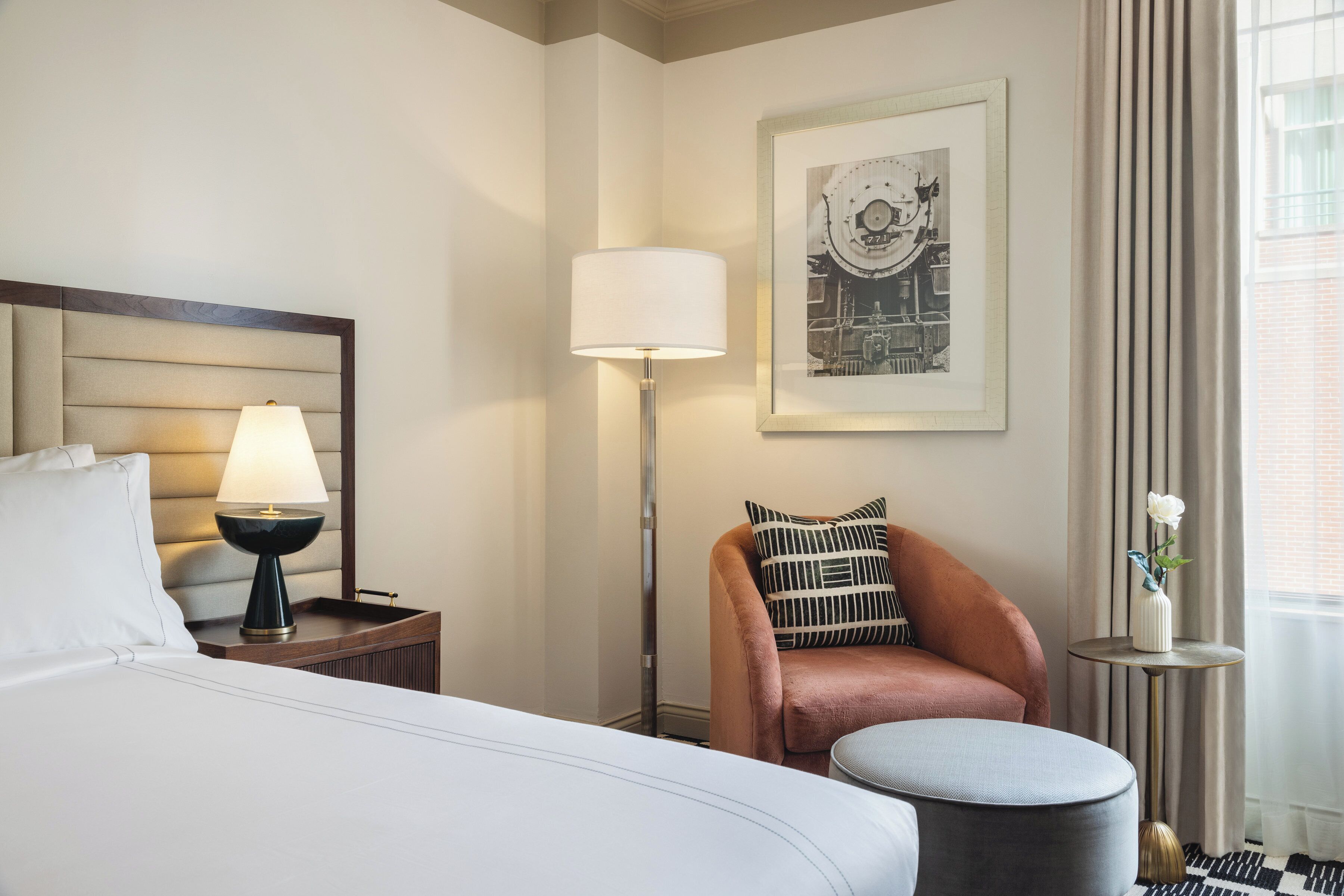 A hotel room with a bed, a nightstand with a lamp, a chair, a small table with a vase, and a framed picture on the wall.