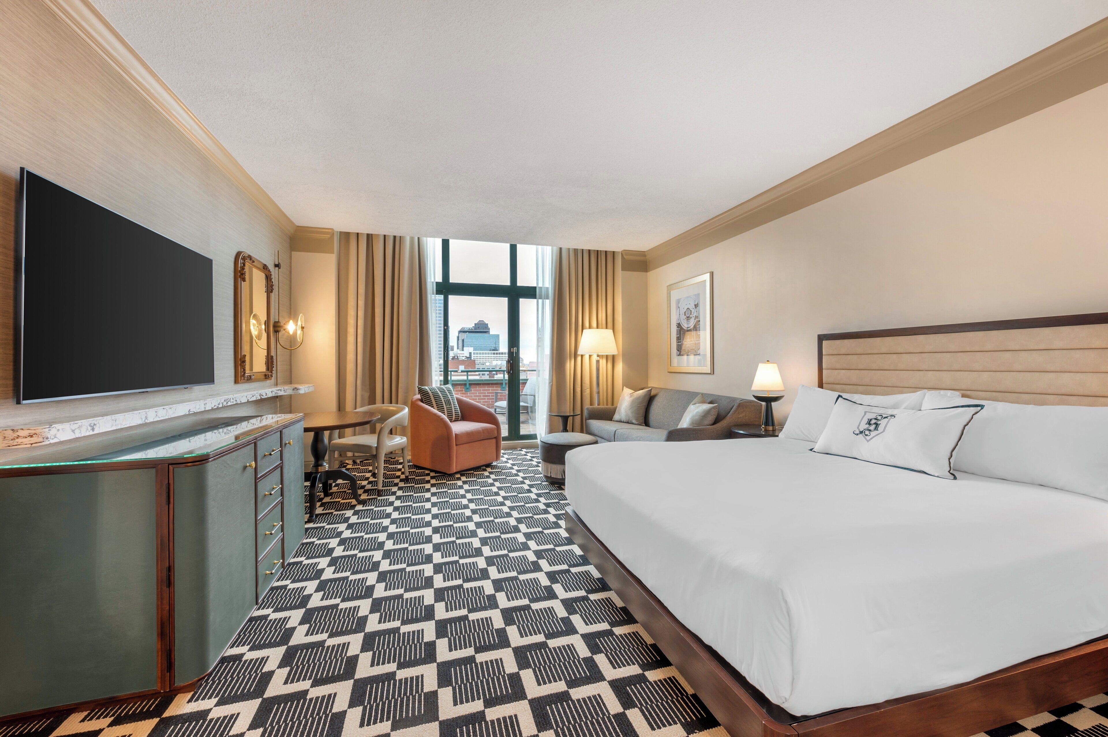 Ett hotellrum med en stor säng, ett skrivbord med en platt-TV, en stol, en soffa och utsikt över staden genom fönstret.