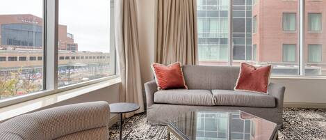 Habitación de hotel con un sofá gris, una mesa redonda pequeña y vistas a un estadio y edificios.