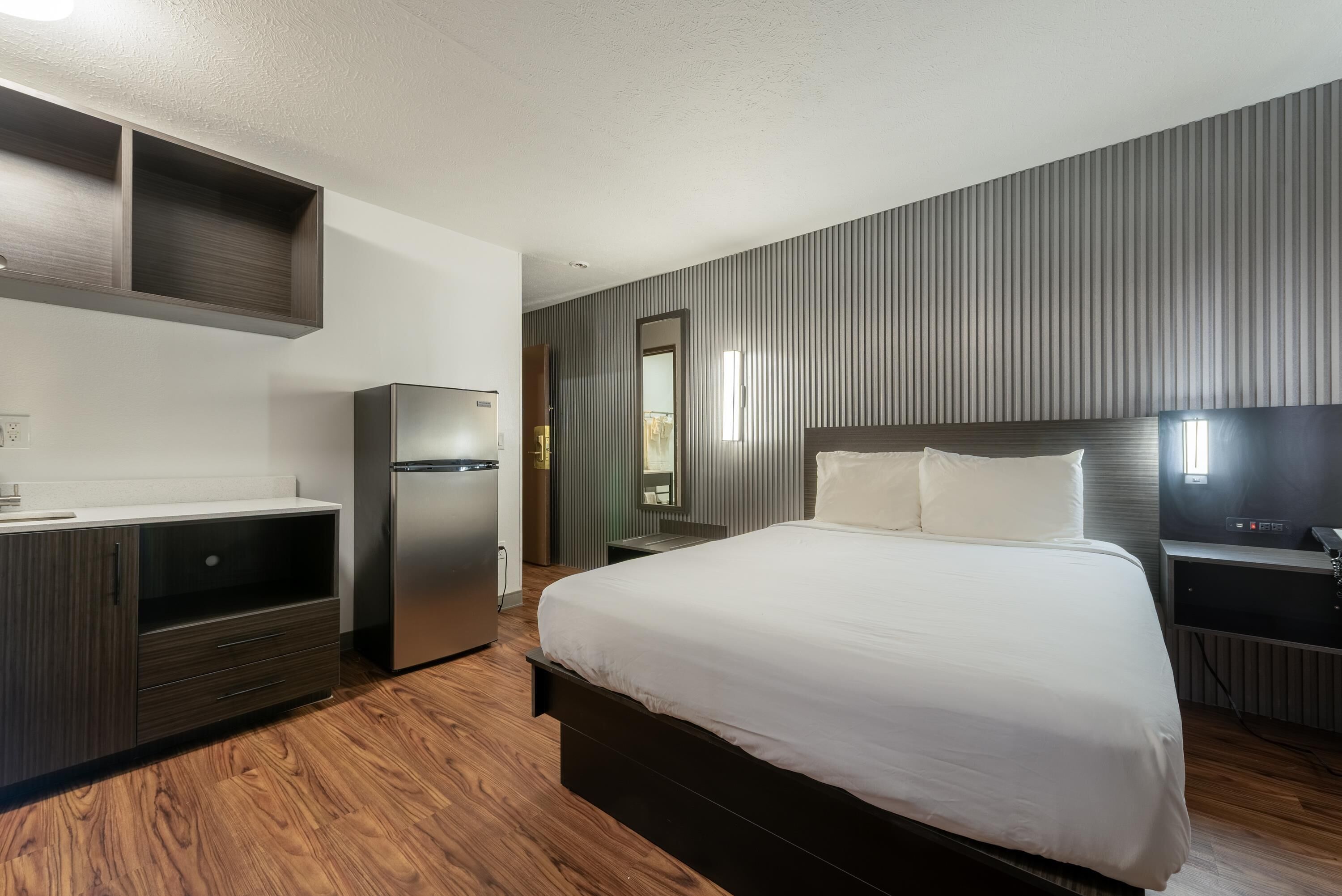 Une chambre d’hôtel moderne équipée d’un lit, d’un bureau intégré, d’un réfrigérateur et d’un téléviseur fixé au mur.