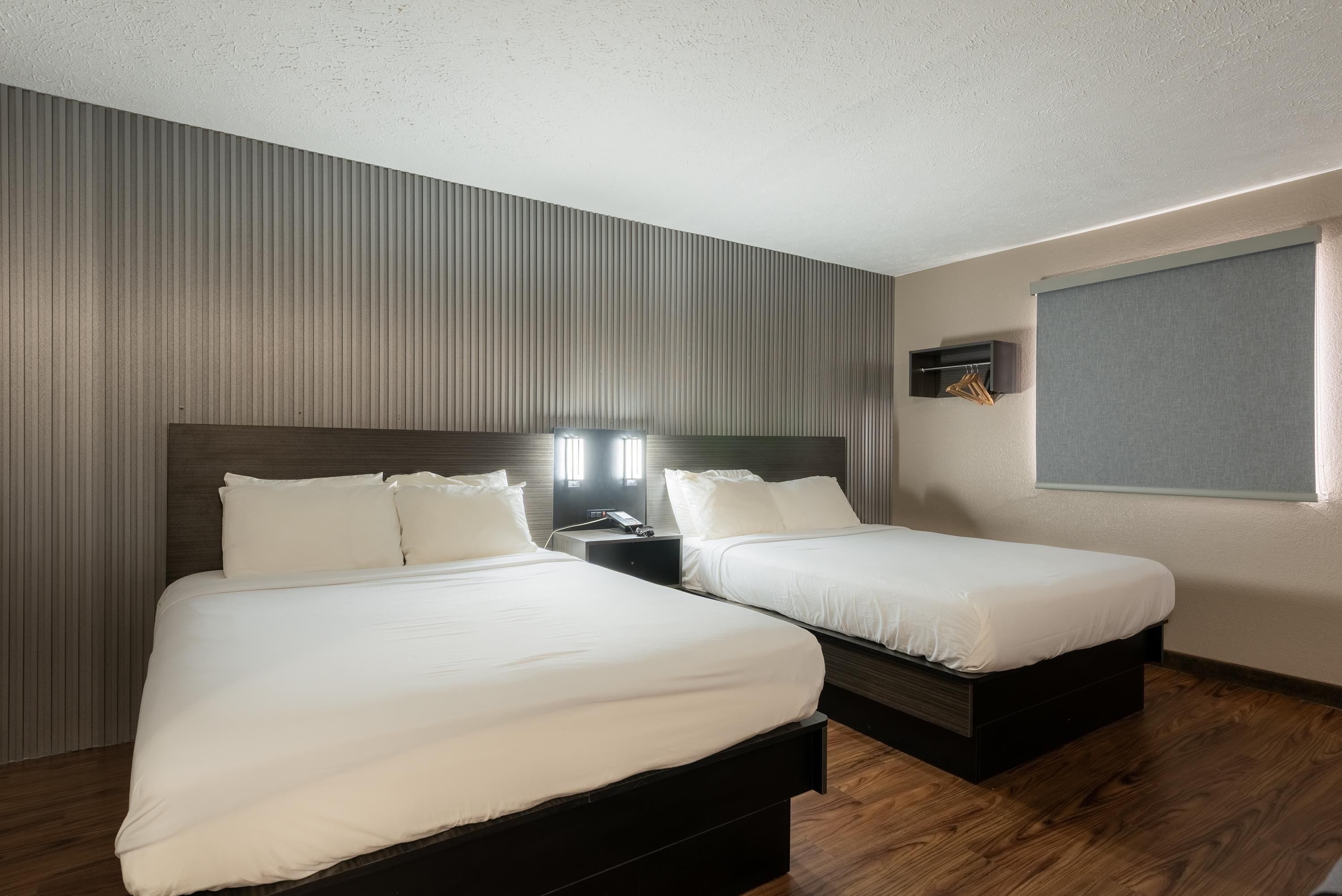 Ein Hotelzimmer mit zwei Betten, einem Nachttisch, einem an der Wand befestigten Regal und einem Fenster mit Jalousien.