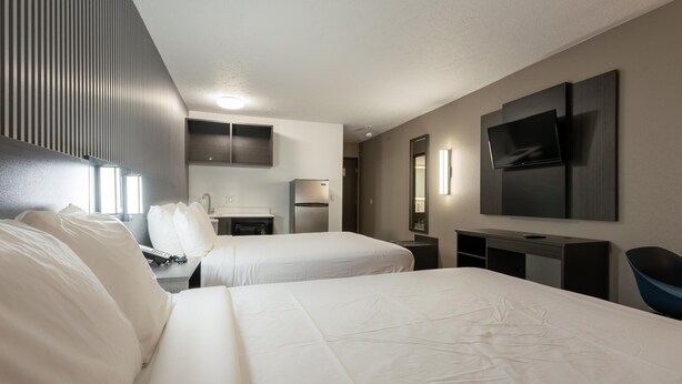 Habitación de hotel con dos camas, un televisor montado en la pared, un escritorio con una silla y una kitchenette pequeña.