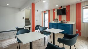 Un área de comedor moderna con mesas blancas y sillas negras, una barra azul con televisor de pantalla plana, y paredes con rayas rojas y blancas.