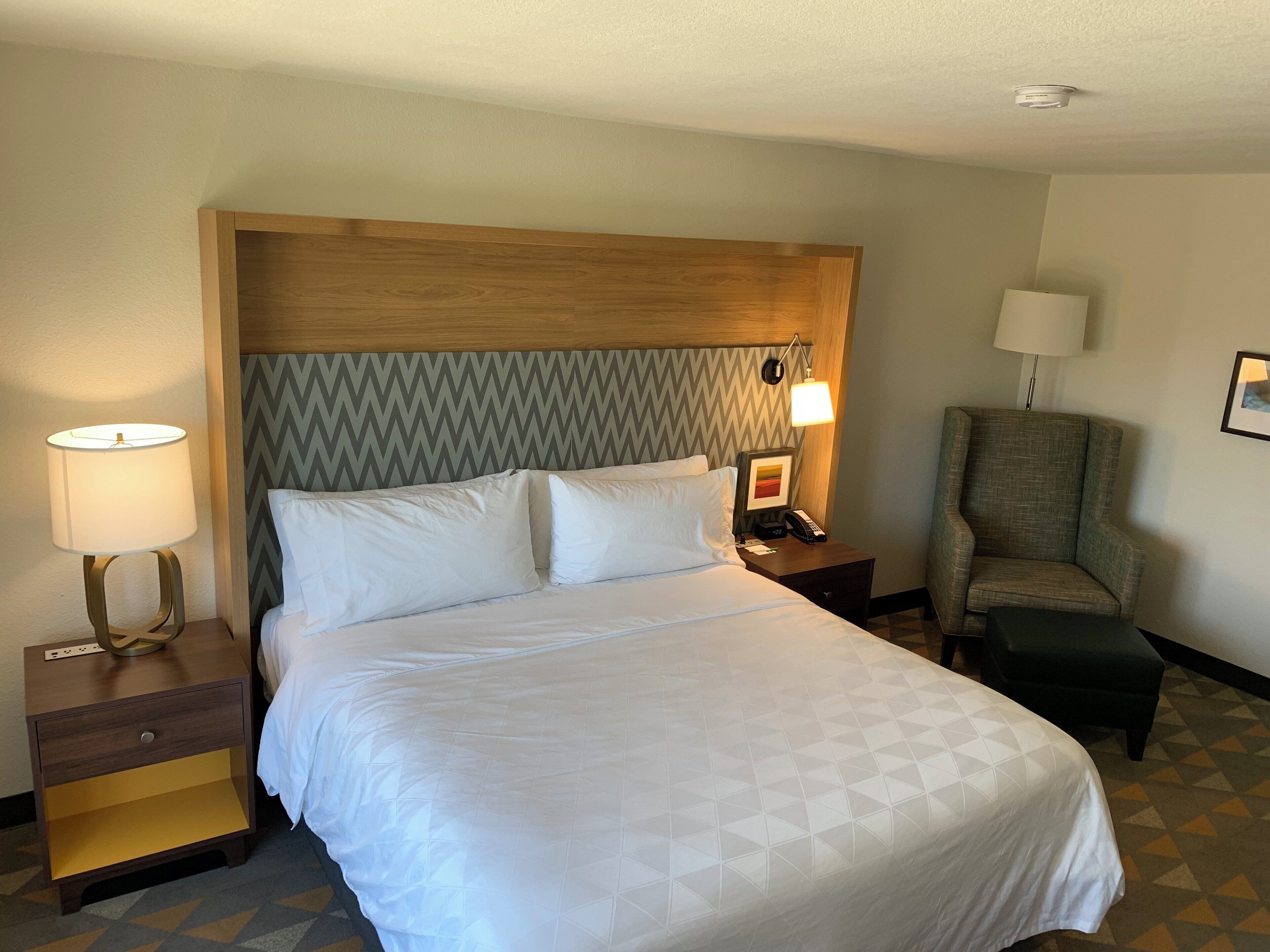 A hotel room with a large bed, a nightstand with a lamp, a chair, and a desk with a telephone.