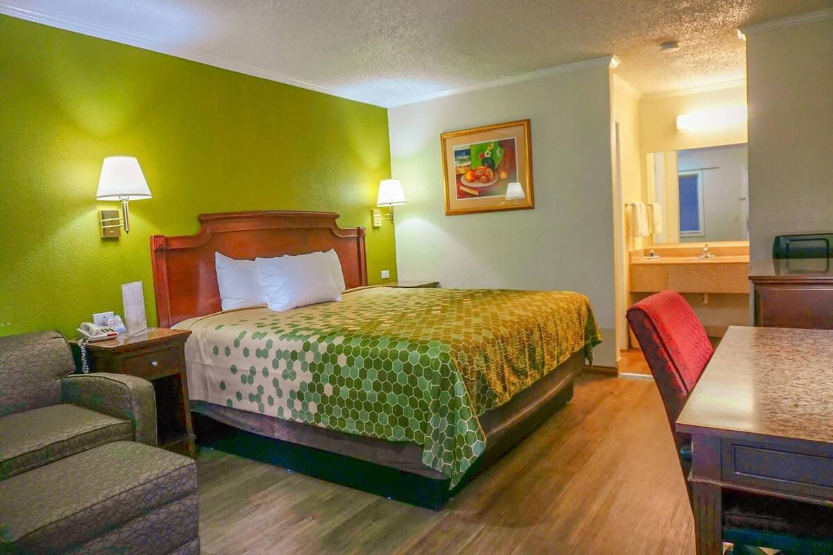 A hotel room with a bed, two bedside lamps, a chair, a table, and a painting on the wall.