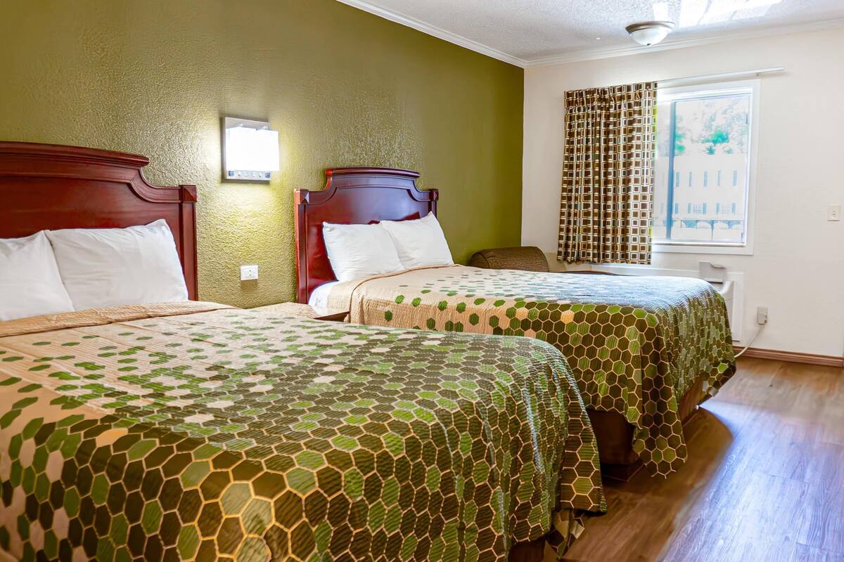 A hotel room with two beds, green patterned bedspreads, wooden headboards, a window with curtains, and a door with a handle.