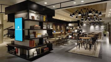A modern hotel lobby with a digital display, bookshelves, dining area, and a staircase.
