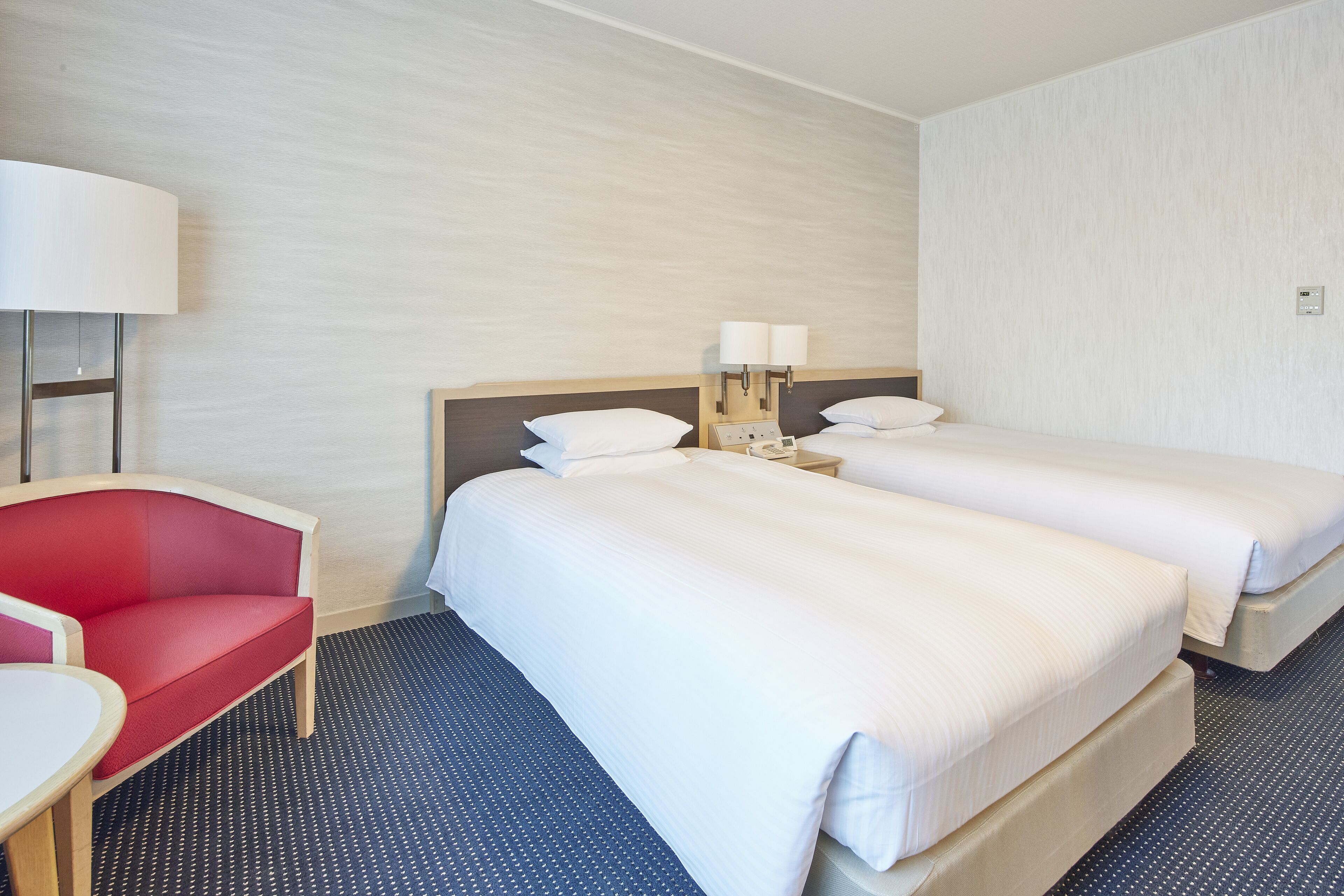 A hotel room with two beds, a red chair, a desk, and a lamp.
