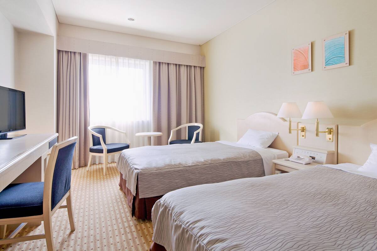 Ein Hotelzimmer mit zwei Betten, einem Schreibtisch mit Fernseher, einem Sessel, einem kleinen Tisch und einem Nachttisch mit Lampen.