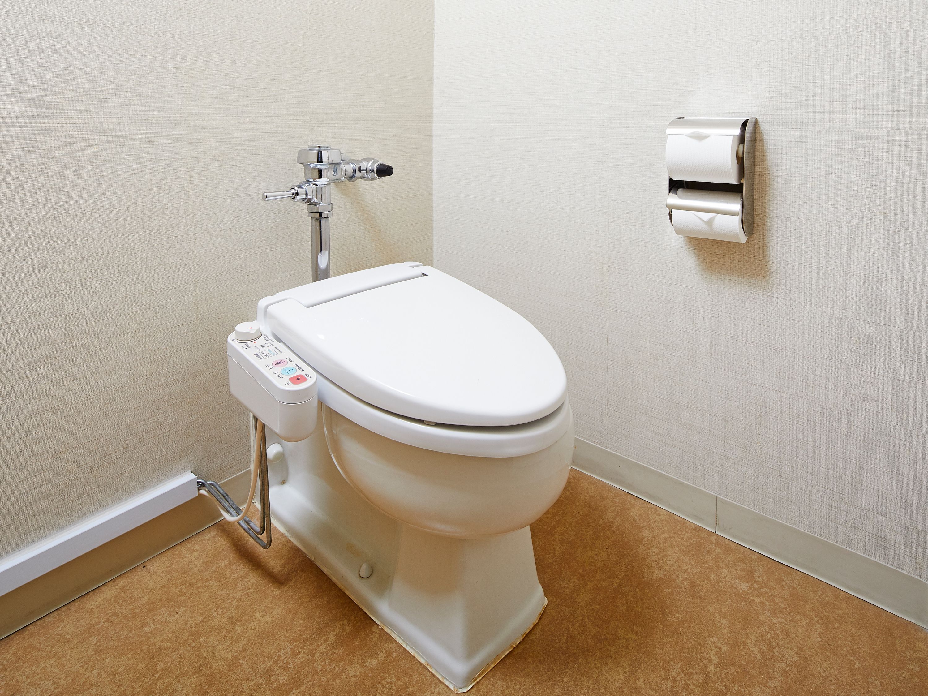 A modern toilet with a control panel, a wall-mounted toilet paper dispenser, and a visible pipe.