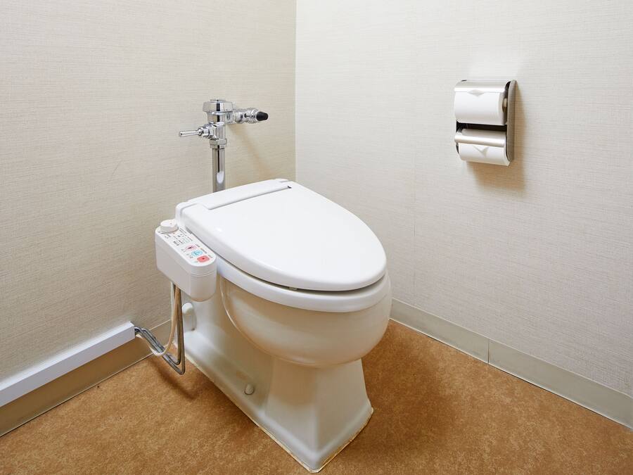 A modern toilet with a control panel, a wall-mounted toilet paper dispenser, and a visible pipe.