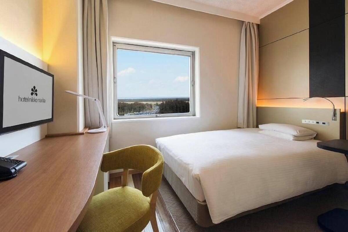 Ein Hotelzimmer mit einem Bett, einem Schreibtisch mit Stuhl, einem Fenster mit Blick und einem am Wand montierten Fernseher, der den Hotelnamen anzeigt.