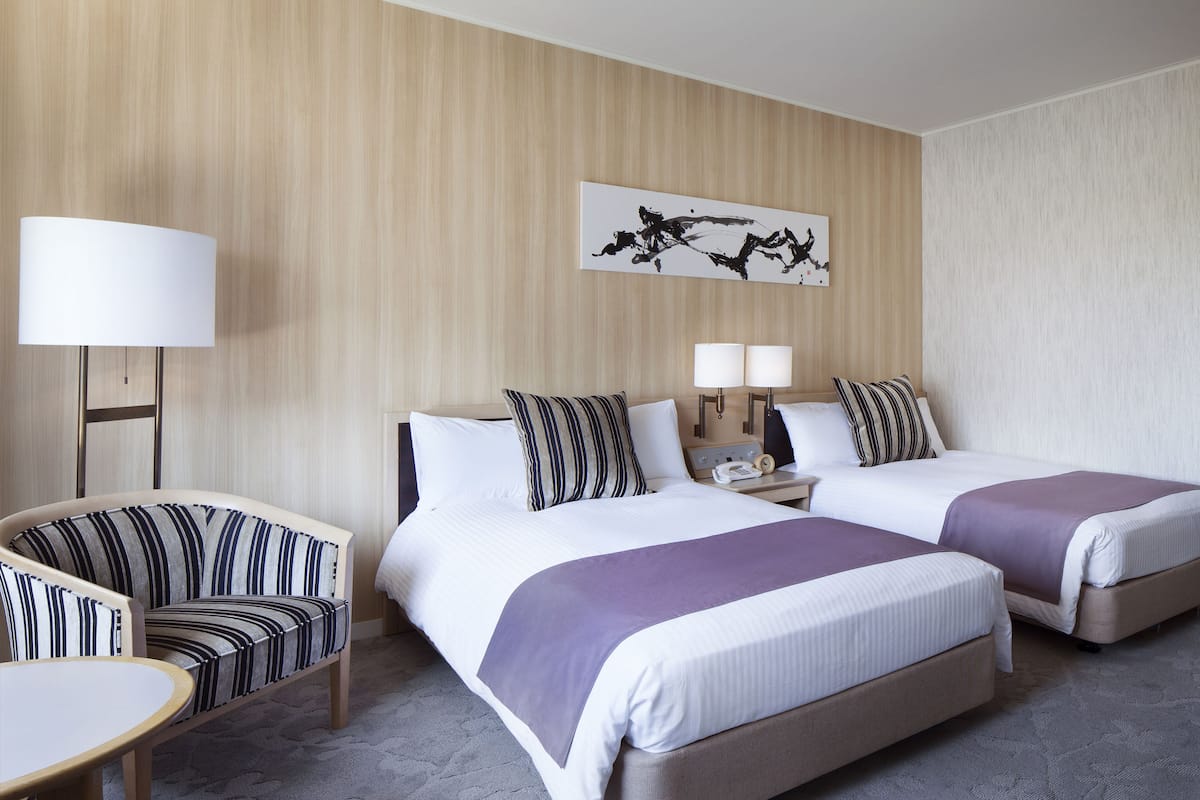 Ein Hotelzimmer mit zwei Betten, einem Sessel, einem Tisch und einer Lampe.