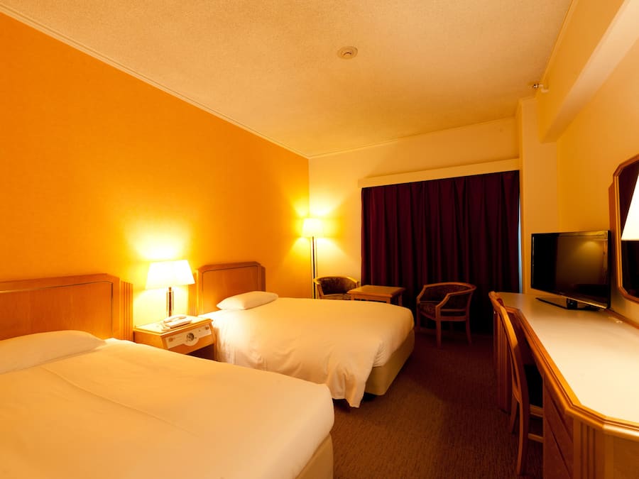 A hotel room with two beds, a desk, a chair, a TV, and a large window with curtains.