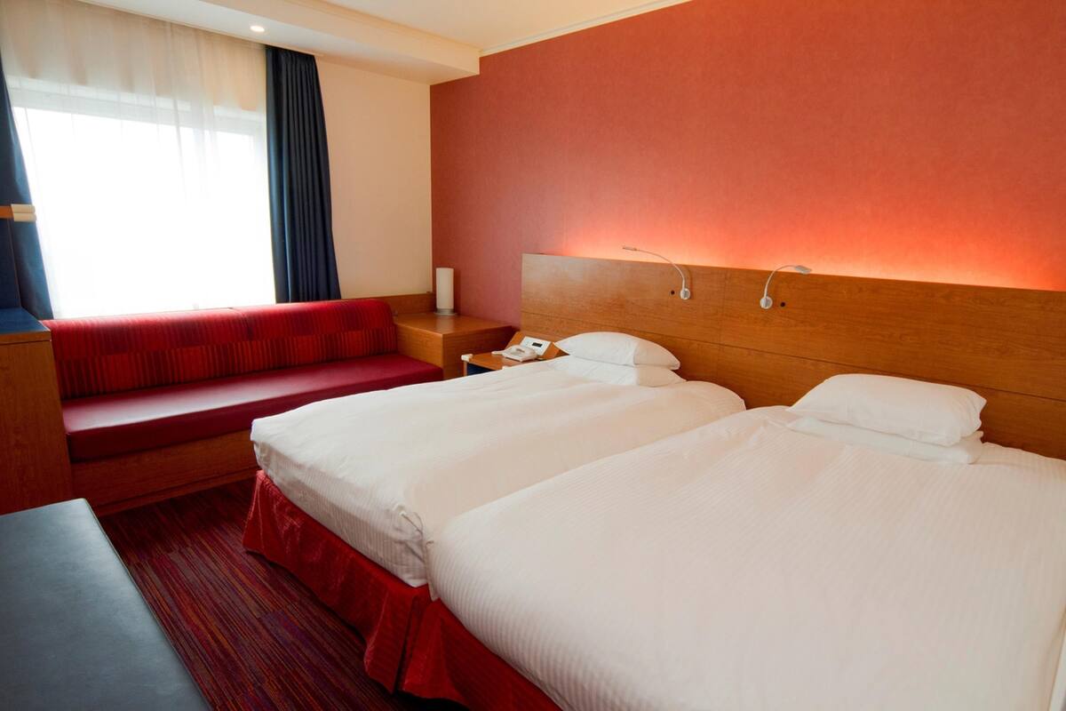 Ein Hotelzimmer mit zwei Betten, einem roten Sofa, einem Holznachttisch und einem Fenster mit Vorhängen.