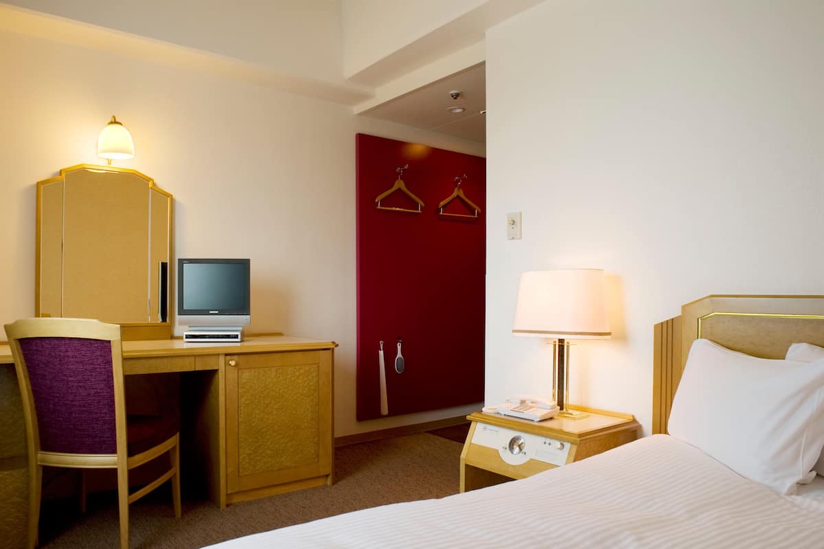 Ein Hotelzimmer mit einem Bett, einem Schreibtisch mit Computer, einem Stuhl, einem Nachttisch mit Lampe und einer roten Tür.