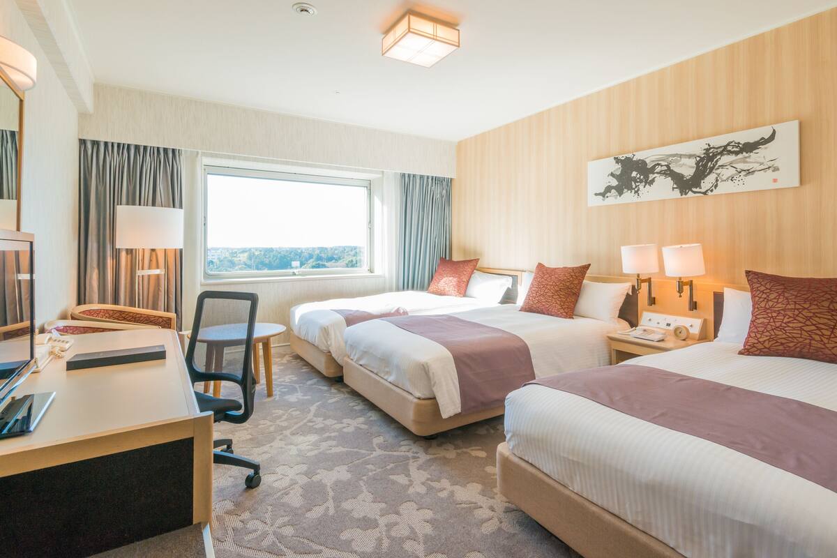 Ein Hotelzimmer mit zwei Betten, einem Schreibtisch mit Stuhl, einer Schreibtischlampe, einem Nachttisch mit Lampe, einem Fenster mit Vorhängen und einem Bild an der Wand.