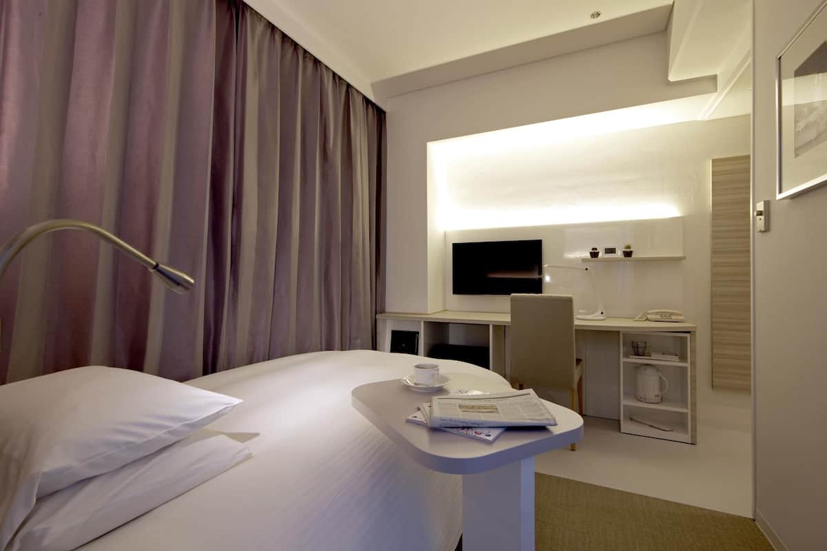 Ein Hotelzimmer mit einem Bett, einem Schreibtisch mit Computer, einem Fernseher und einer Kaffeetasse auf dem Nachttisch.