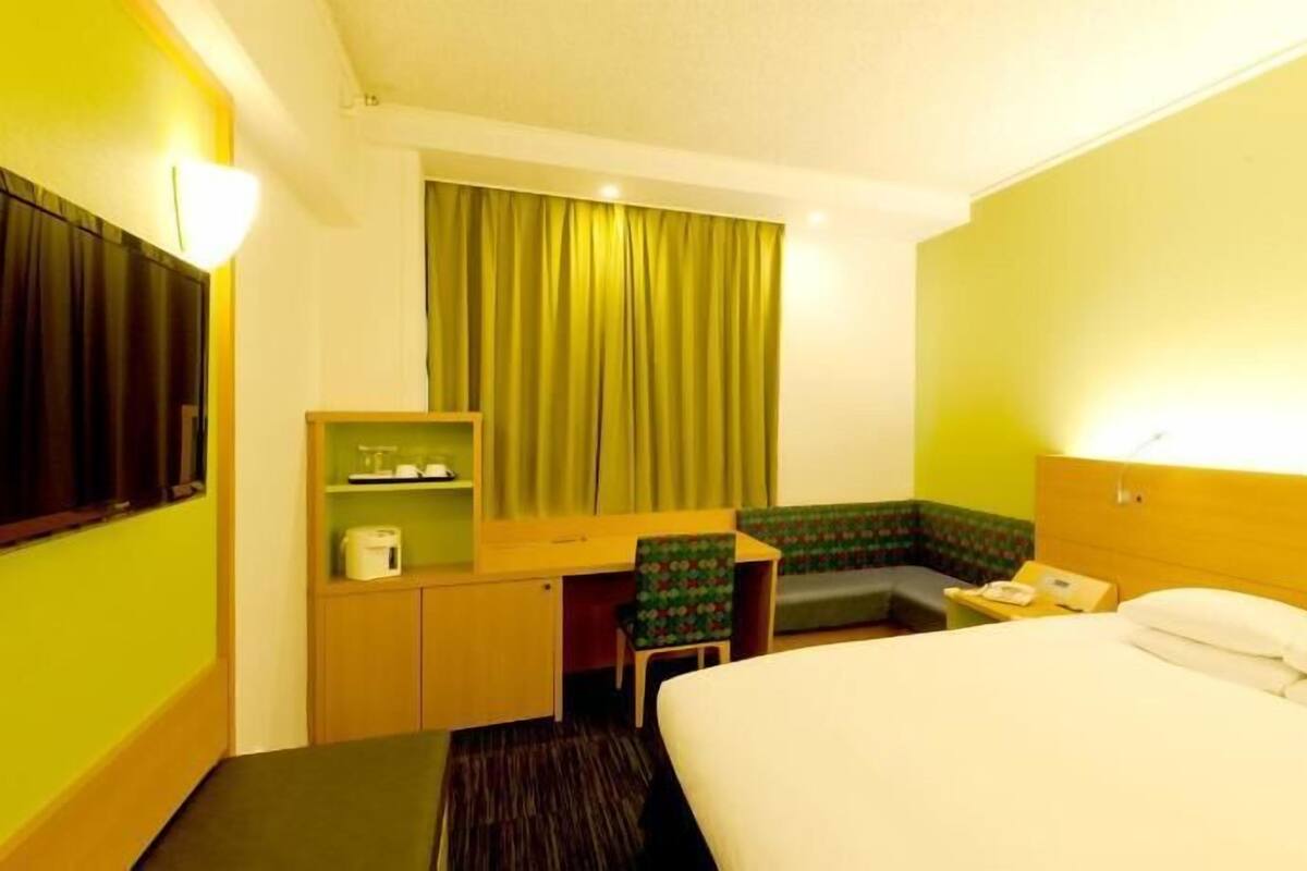 Ein Hotelzimmer mit einem Bett, einem Schreibtisch, einem Stuhl, einem kleinen Tisch, einem Telefon und einem Fenster mit Vorhängen.