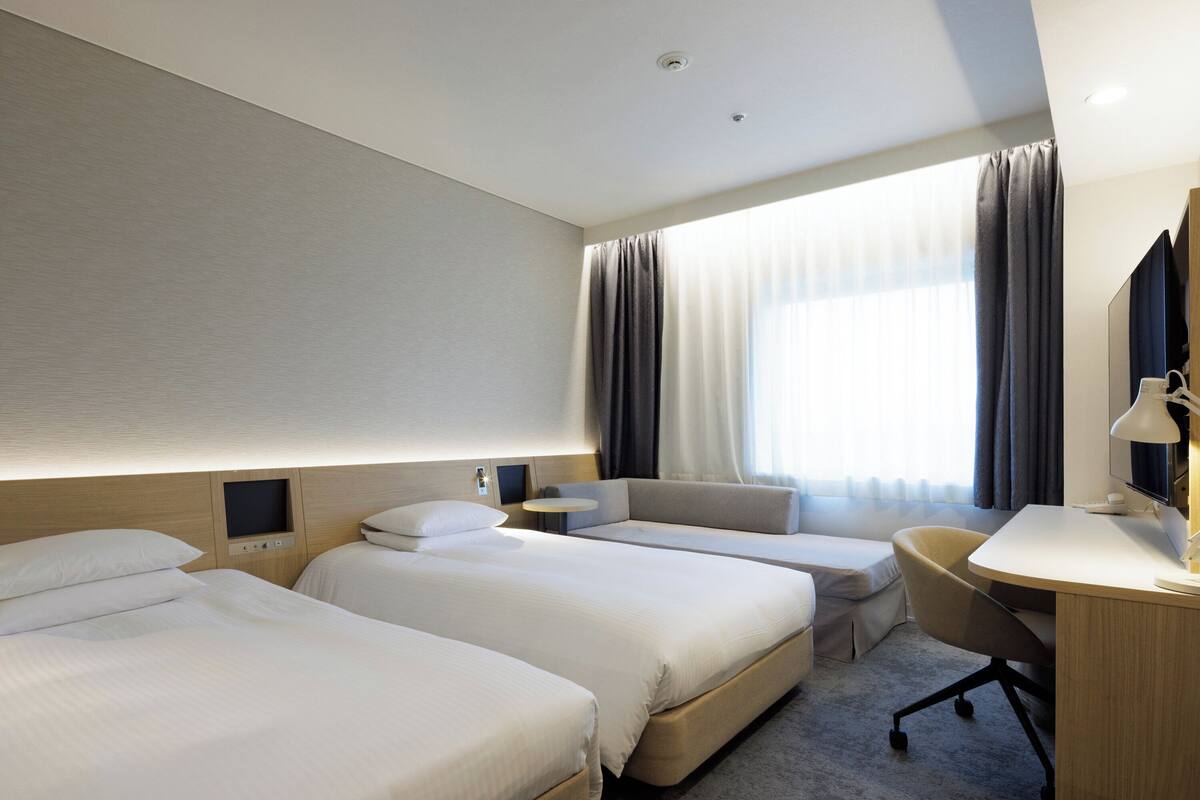 Ein Hotelzimmer mit zwei Betten, einem Schreibtisch, einem Stuhl und einem großen Fenster mit Vorhängen.