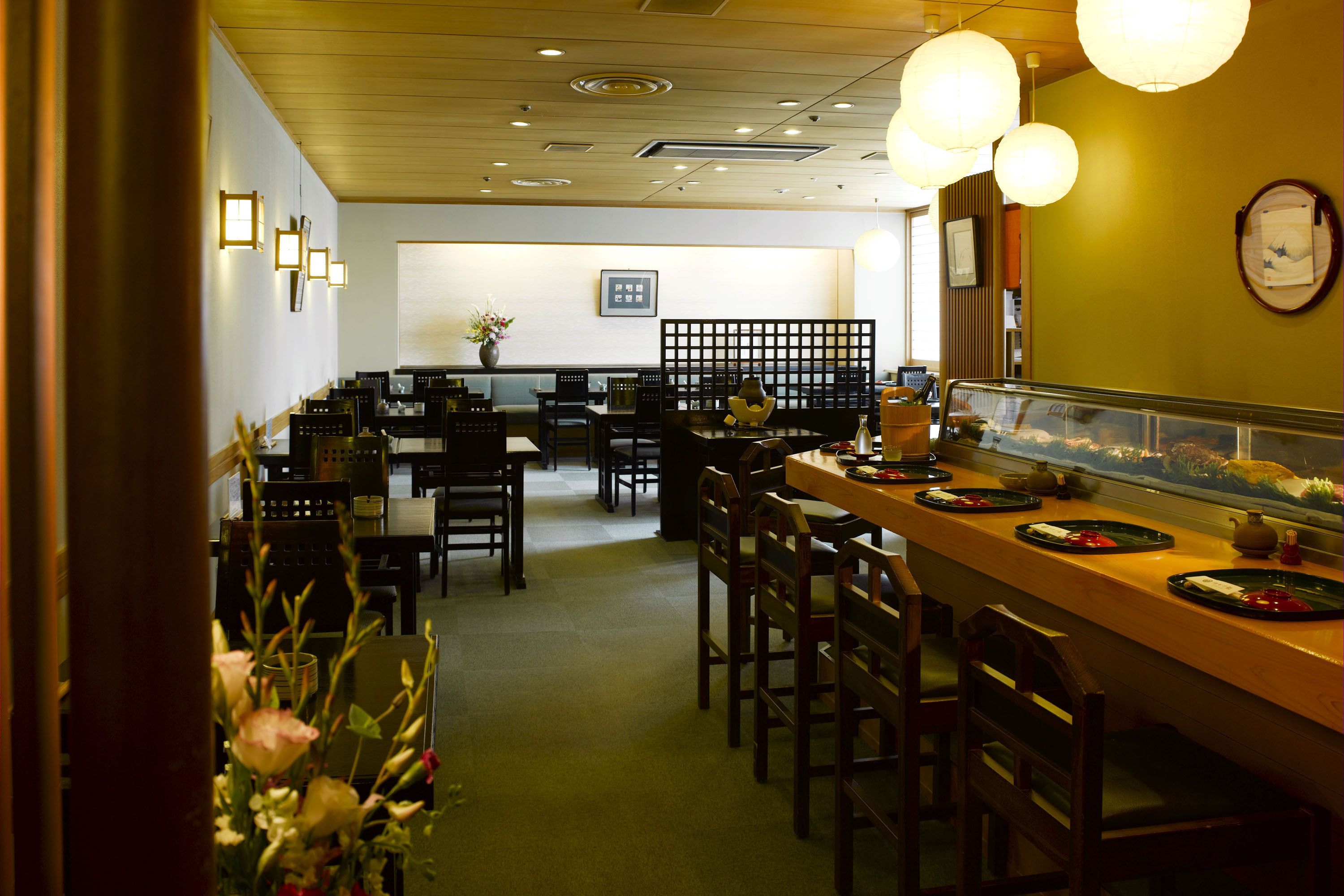 A restaurant with a wooden bar, multiple tables, and chairs. There are hanging lights and a clock on the wall.