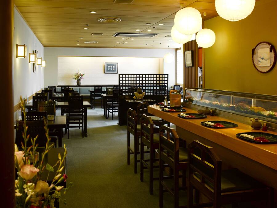 A restaurant with a wooden bar, multiple tables, and chairs. There are hanging lights and a clock on the wall.