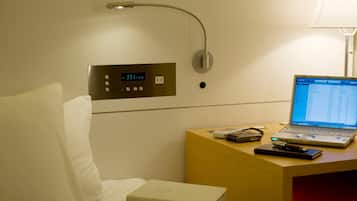 Habitación de hotel con cama, mesita de noche con una laptop, reloj digital y panel de control en la pared.