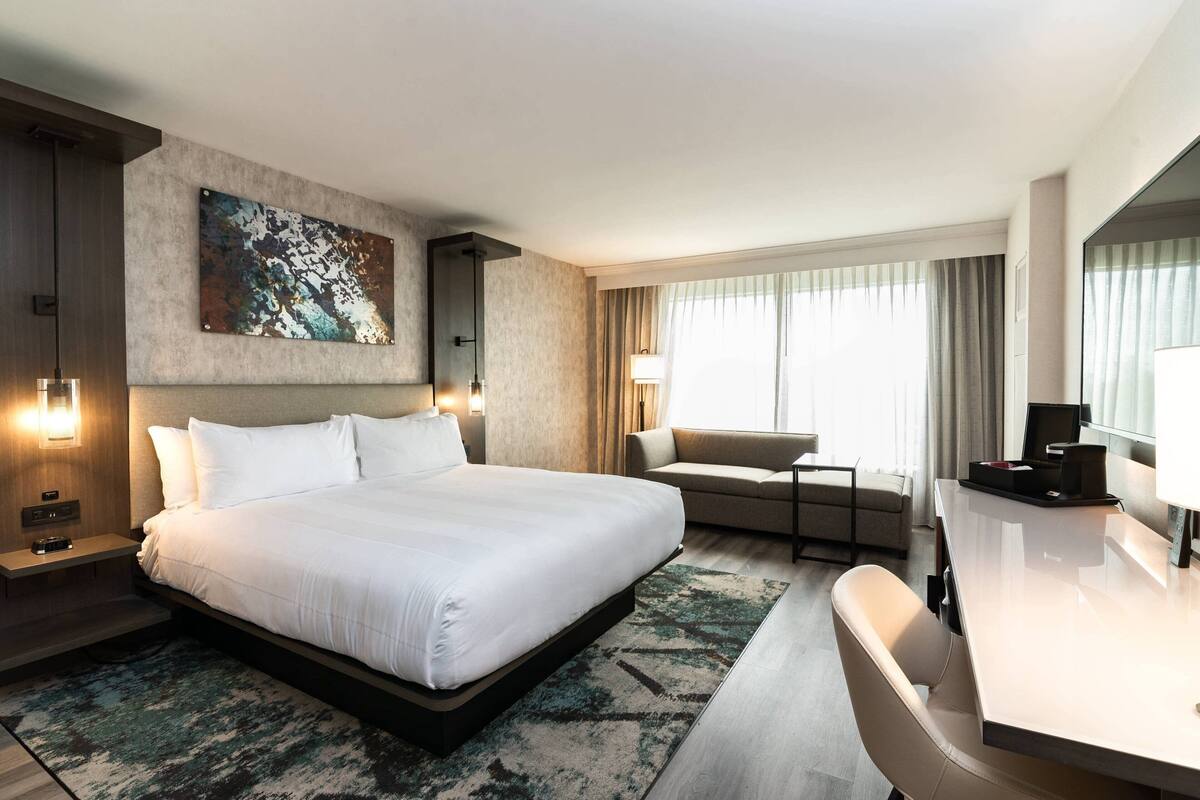 A modern hotel room with a large bed, a desk, a sofa, and a large window with curtains.