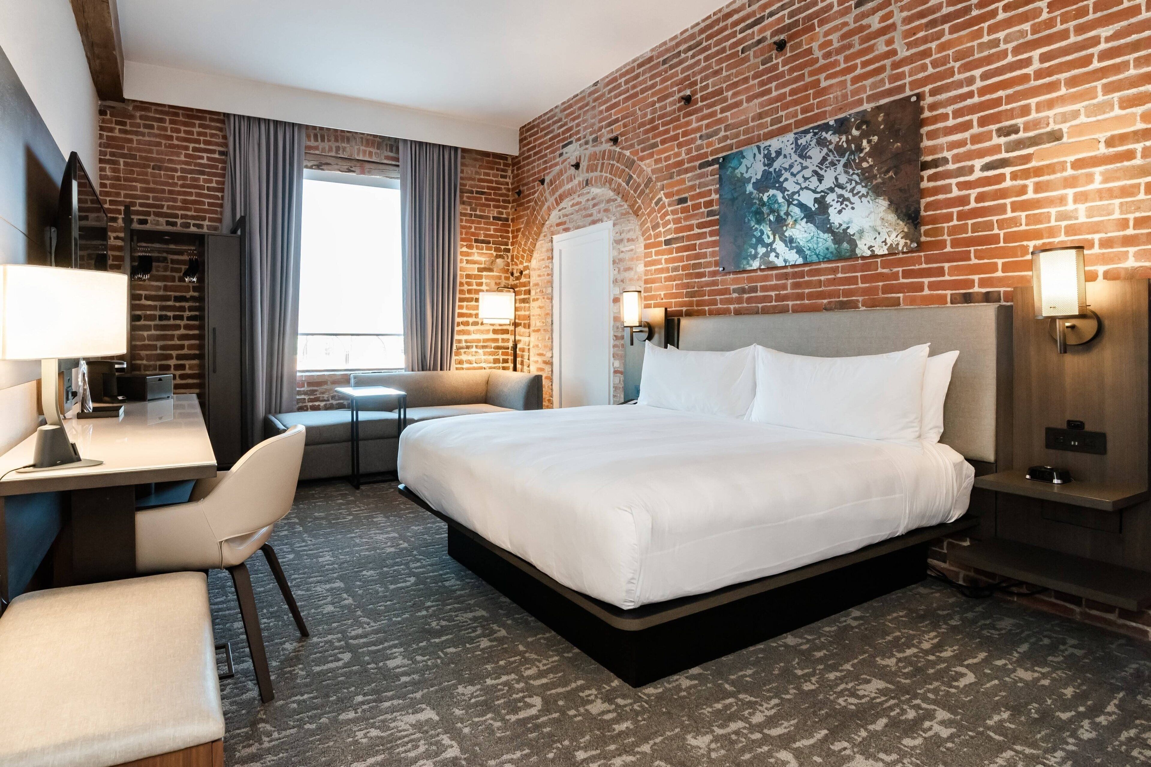 A hotel room with a brick wall, a large bed, a desk, and a chair.