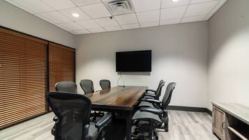 A conference room with a wooden table, black office chairs, a mounted flat-screen TV, and a window with blinds.
