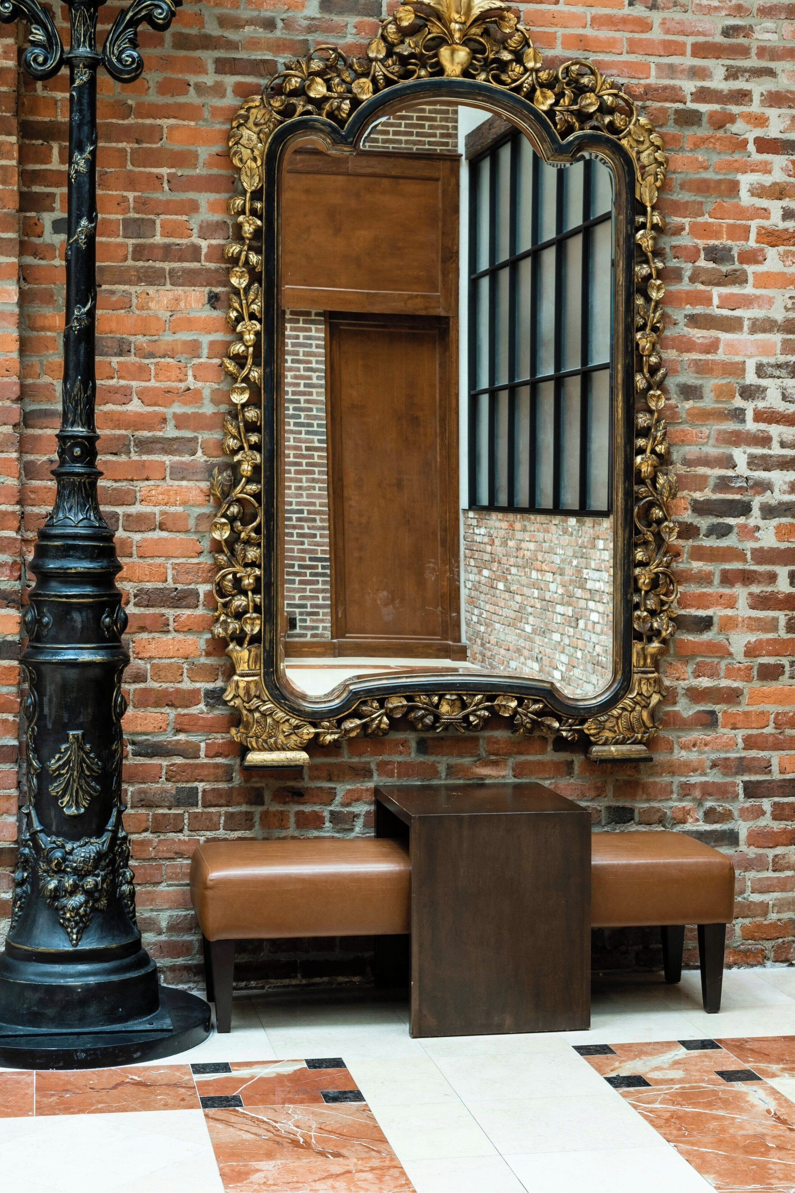 A mirror with an ornate gold frame, a wooden bench, and a black column with a decorative top.