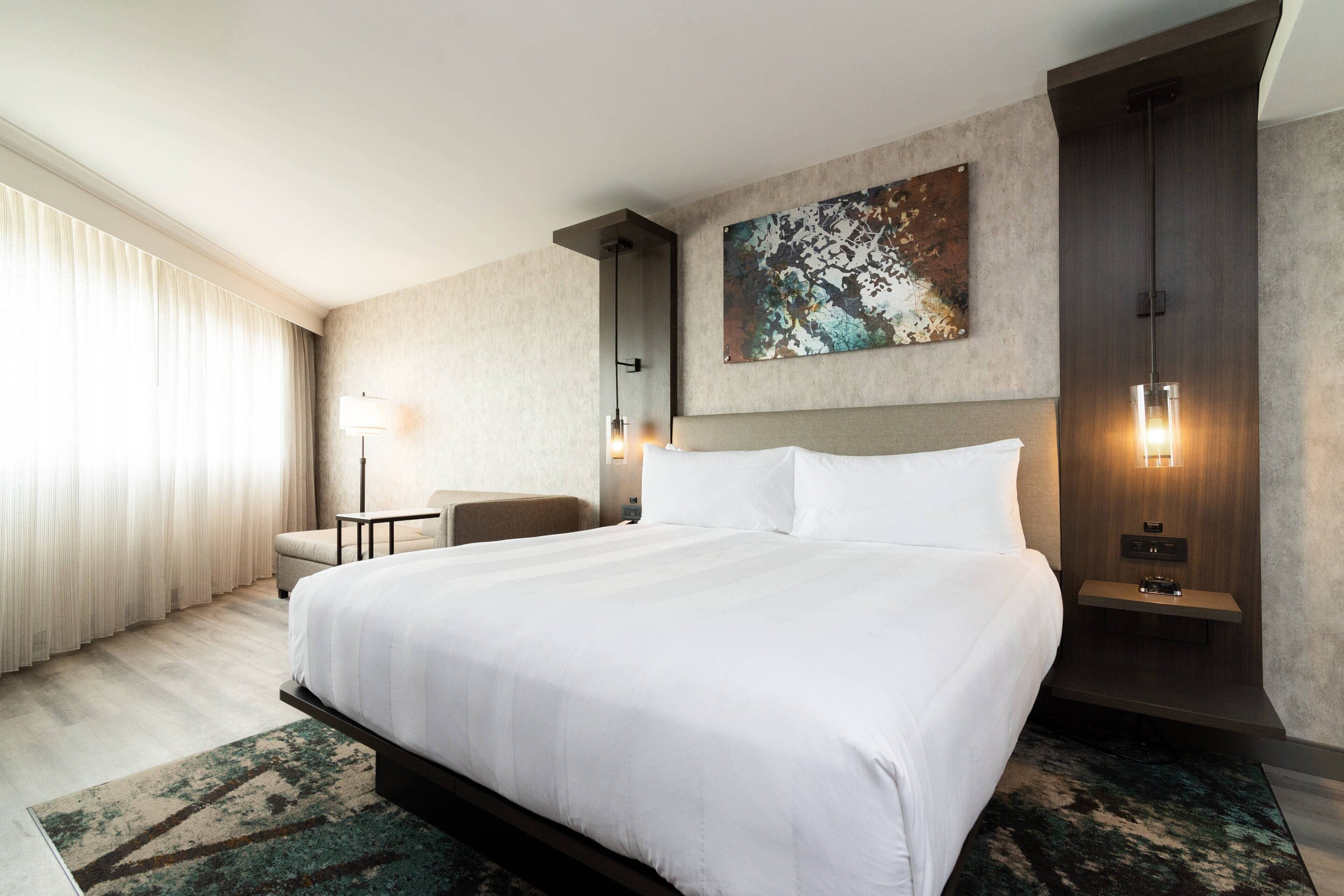 Una habitación de hotel con una cama grande, un escritorio, una silla, un cuadro en la pared y una mesita de noche con lámpara.