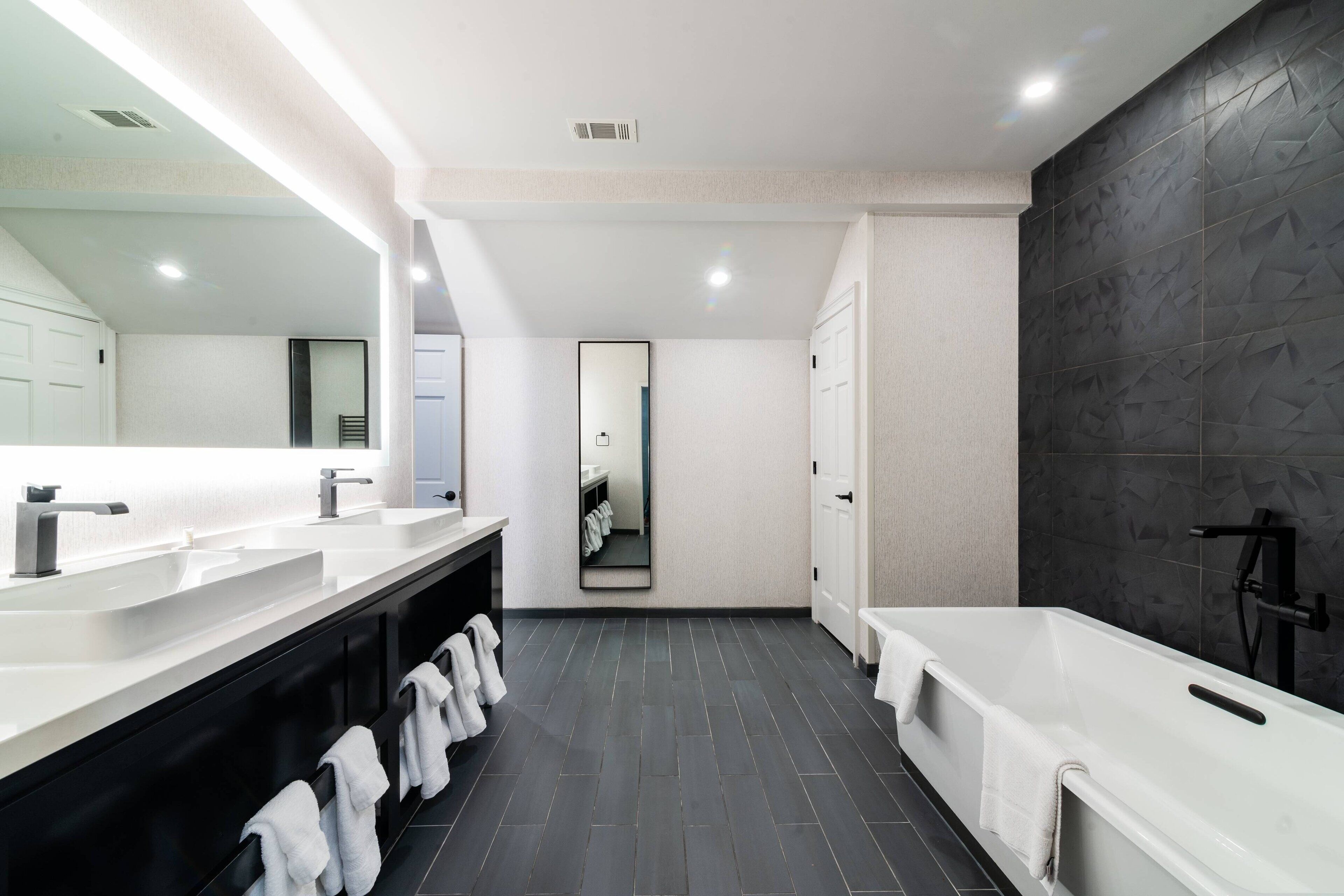 A modern bathroom with dual sinks, a large mirror, a bathtub, and a dark tiled floor.