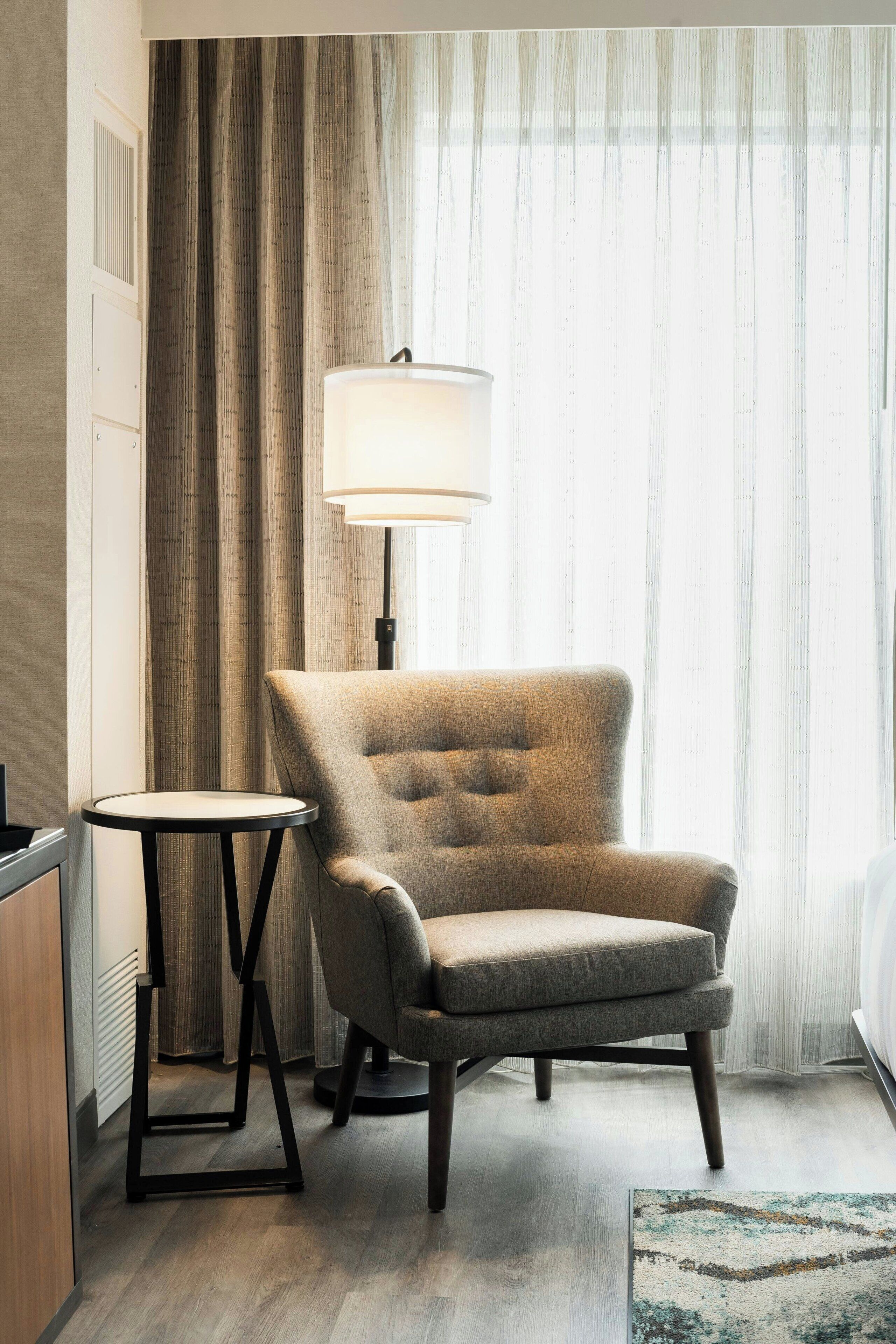 A hotel room with a single armchair, a small round table, a lamp, and a bed.