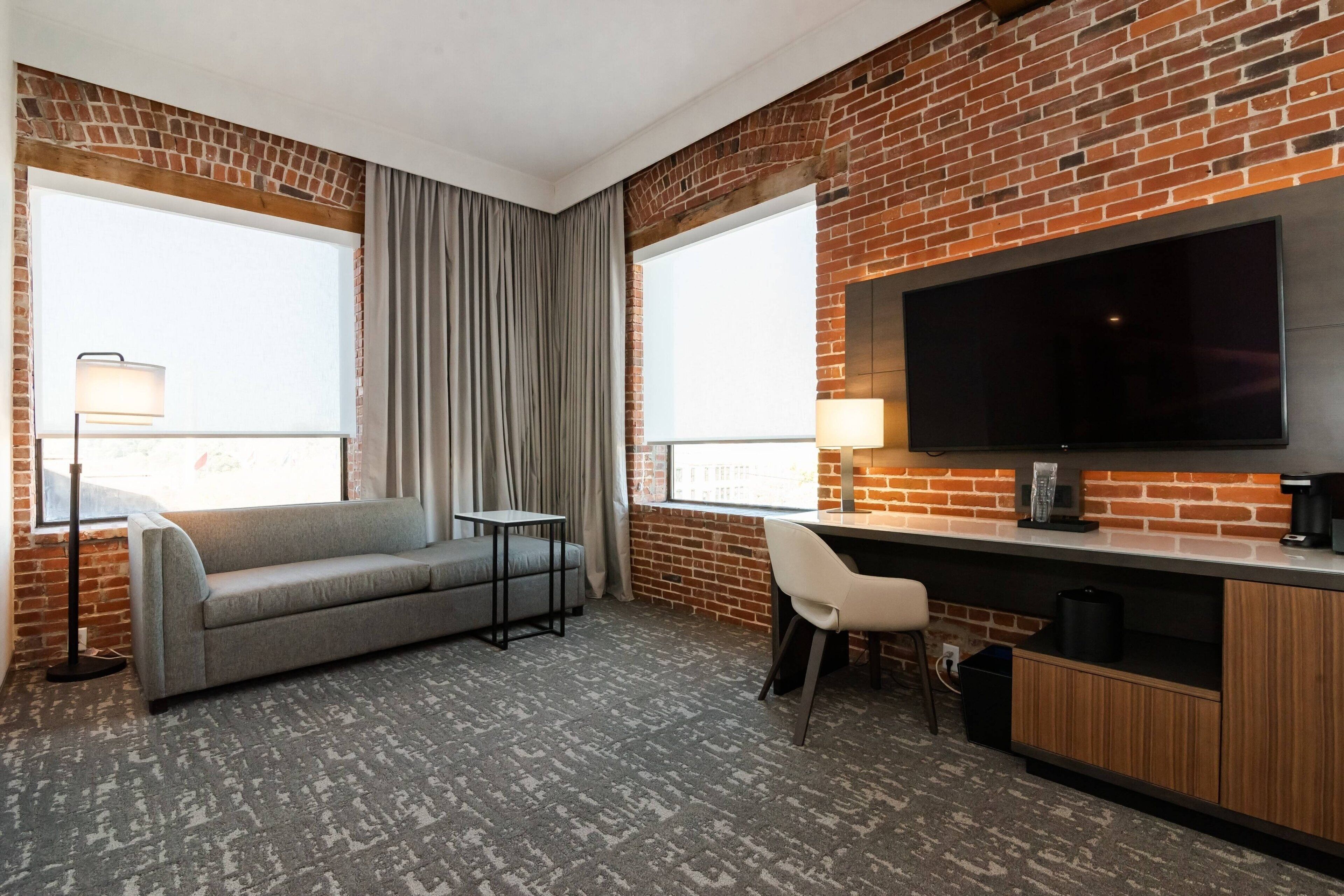 Habitación de hotel con pared de ladrillo, un televisor de pantalla plana, un sofá gris, una mesita, una silla y un escritorio con una cafetera.