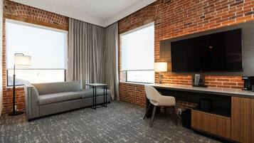 Habitación de hotel con pared de ladrillo, un televisor de pantalla plana, un sofá gris, una mesita, una silla y un escritorio con una cafetera.
