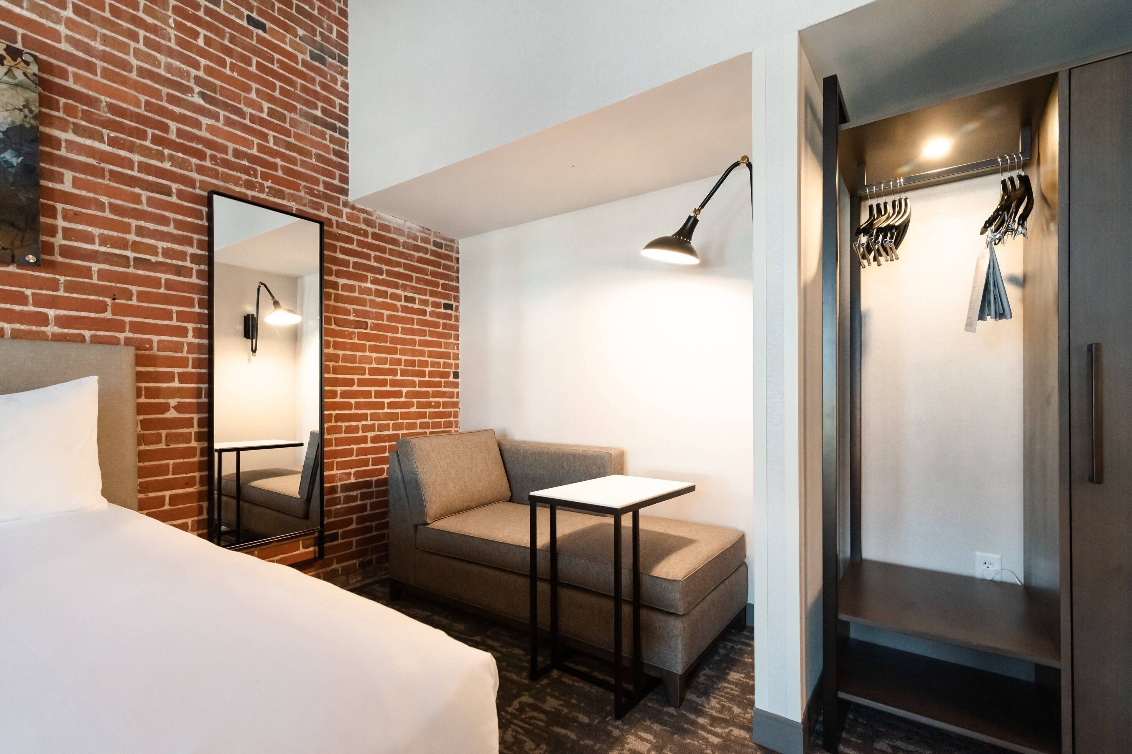 A hotel room with a brick wall, a sofa, a small table, a mirror, a lamp, and a wardrobe with hanging clothes.