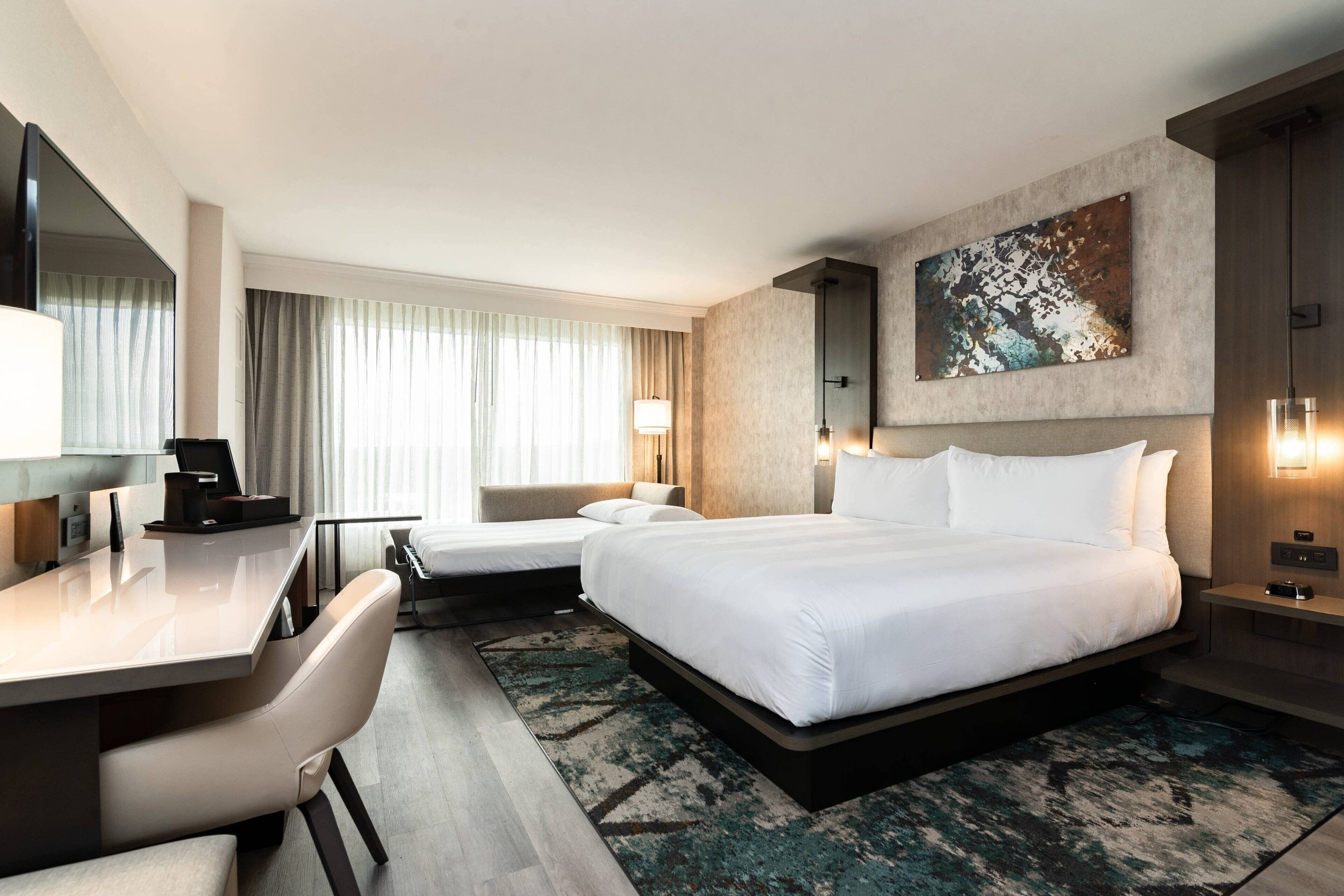 A modern hotel room with a large bed, a desk, and a chair. There is a large window with curtains, a painting on the wall, and a lamp on the nightstand.