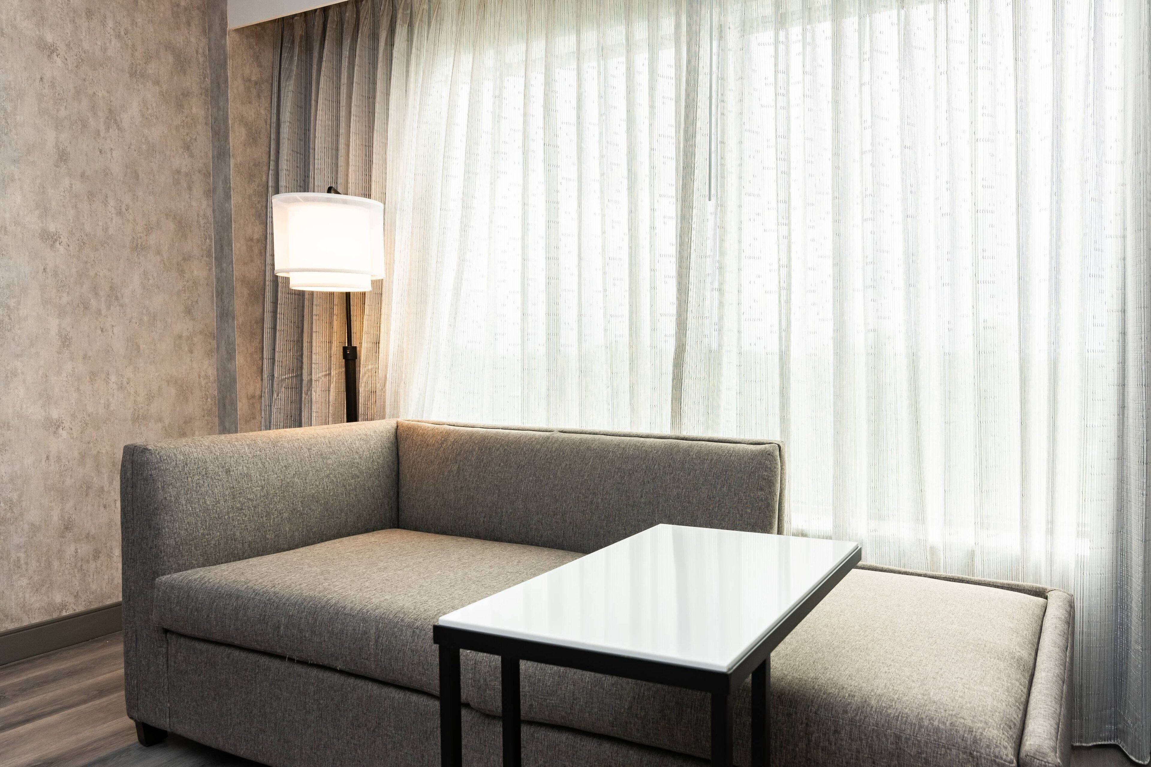 Et moderne hotelværelse med en grå sofa, et lille hvidt bord og en gulvlampe.