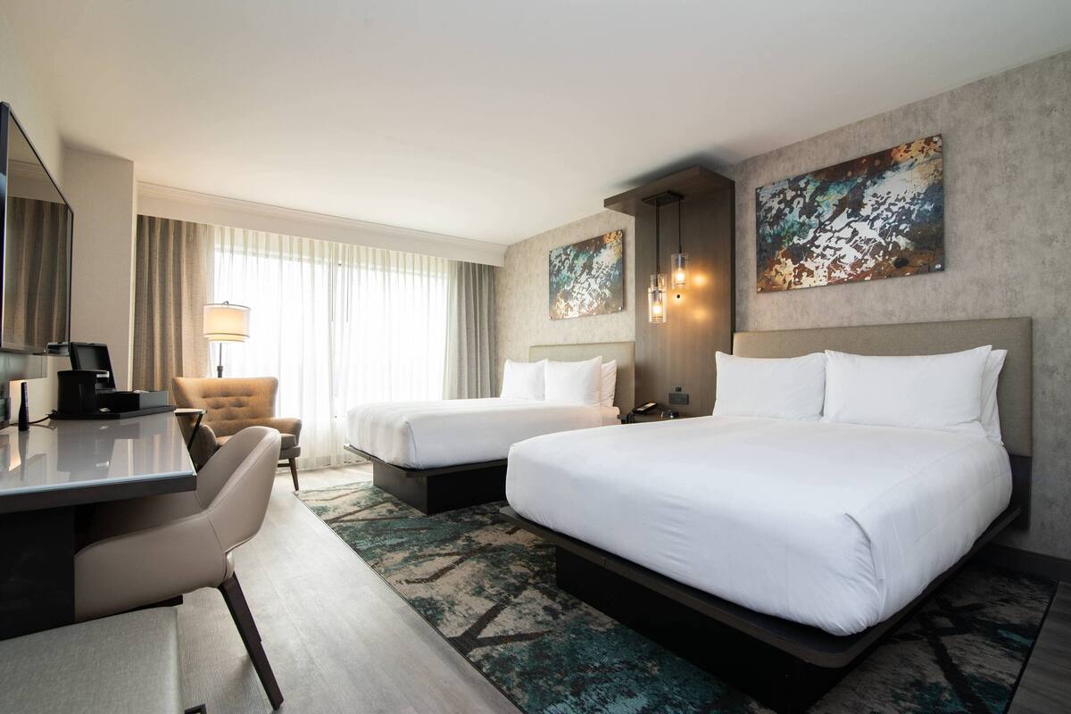 A hotel room with two beds, a desk, a chair, and a large window with curtains.