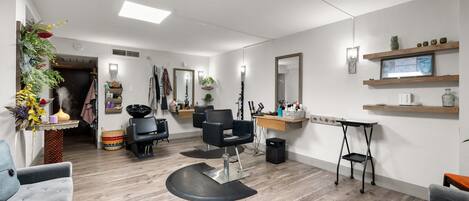 A modern salon with a central black salon chair, wooden flooring, and a mirror above a shelf with decorative items.