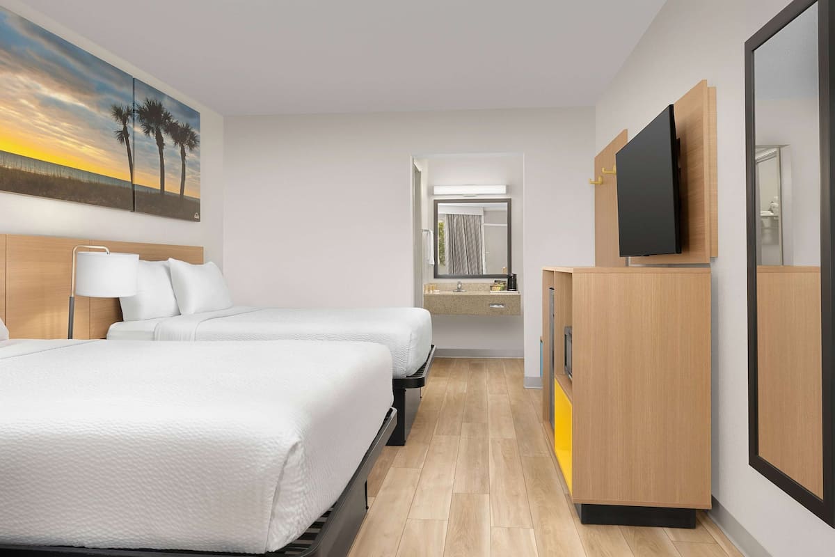 Habitación de hotel con dos camas, un televisor montado en la pared, baño con espejo y un cuadro de palmeras.