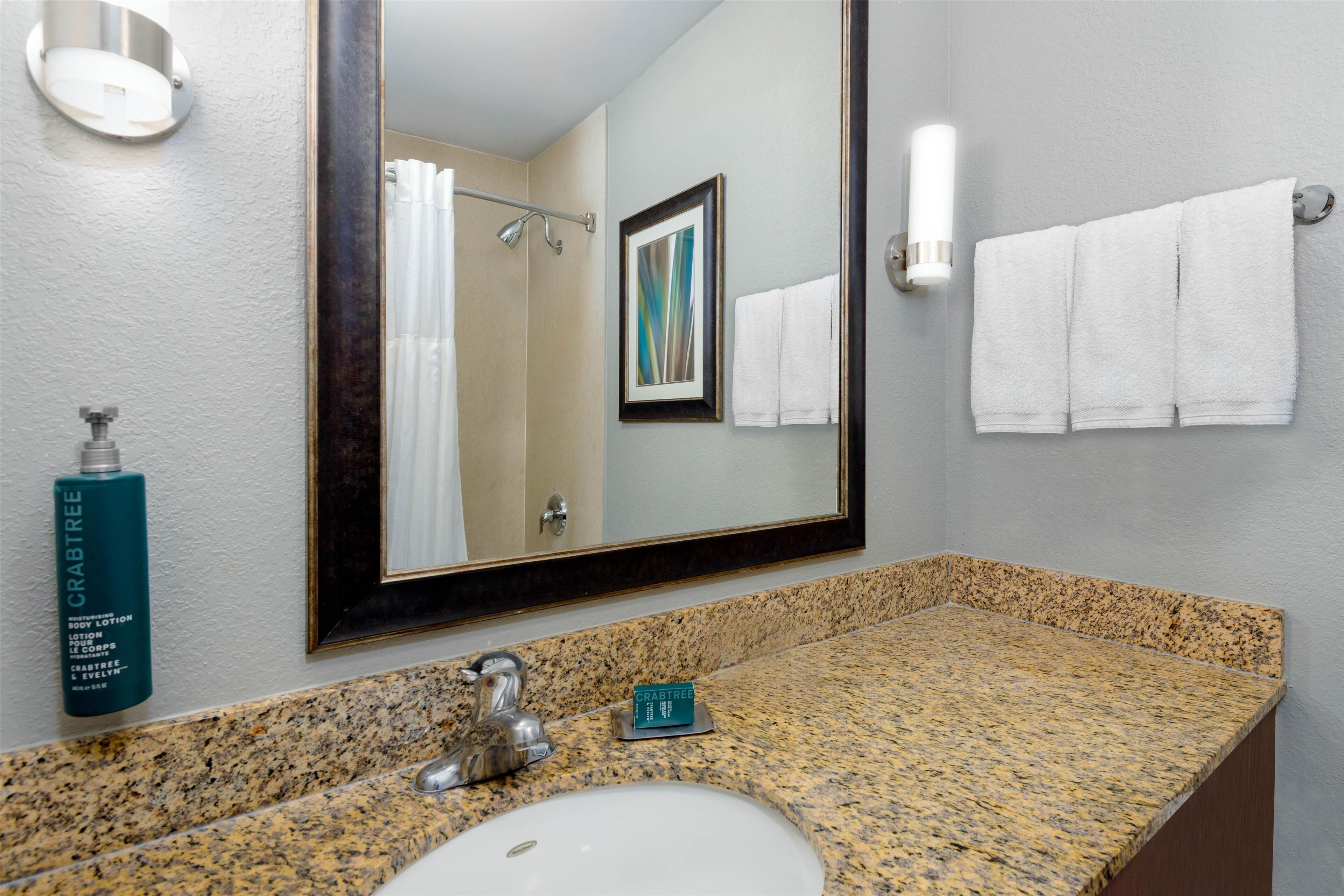 A bathroom with a granite countertop, a sink, a mirror, a framed picture, and a bottle of Crabtree body lotion.
