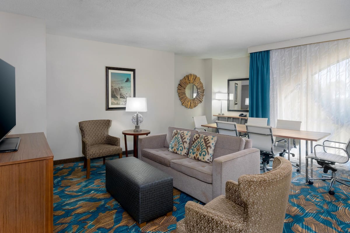 Habitación de hotel con sofá, sillón, reposapiés, escritorio y una ventana grande con cortinas.