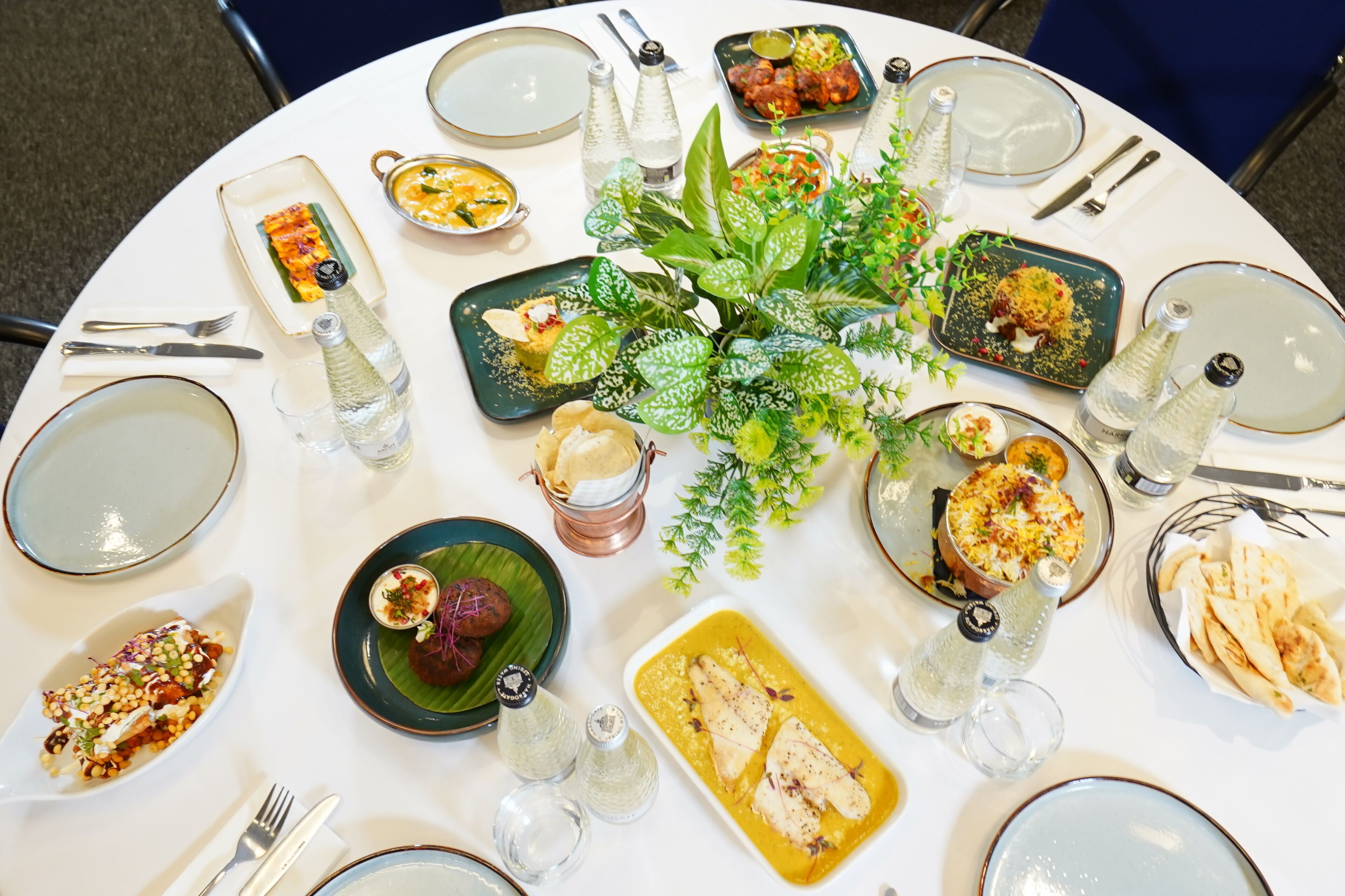 Una mesa redonda de comedor con diversos platos, utensilios y plantas decorativas.